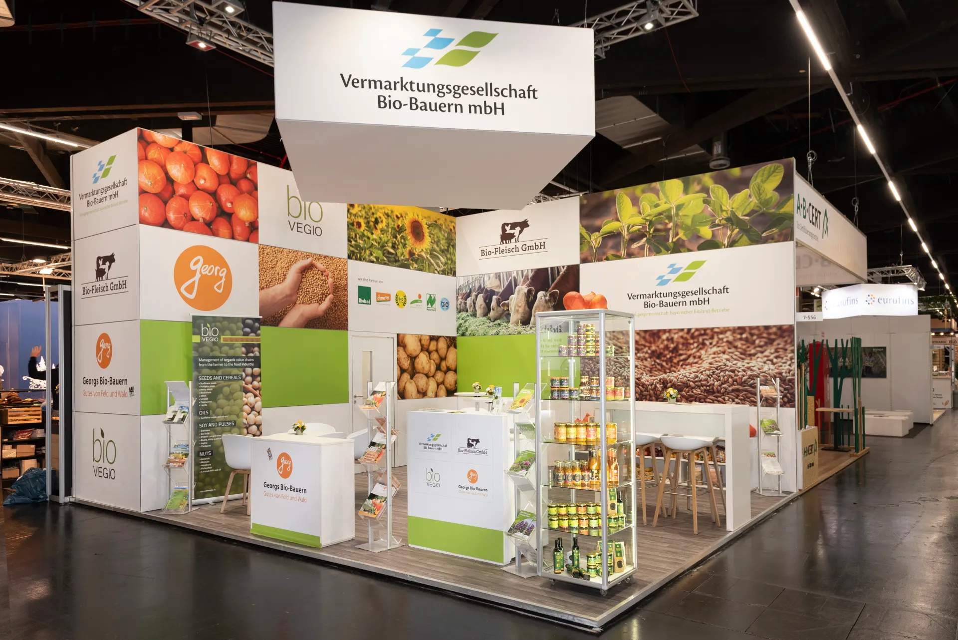 Präsentationsstand mit Glasvitrine voller Bio-Lebensmittel und Sitzgelegenheiten, Messebau der Vermarktungsgesellschaft Bio-Bauern mbH auf der Biofach Messe Nürnberg 2025.