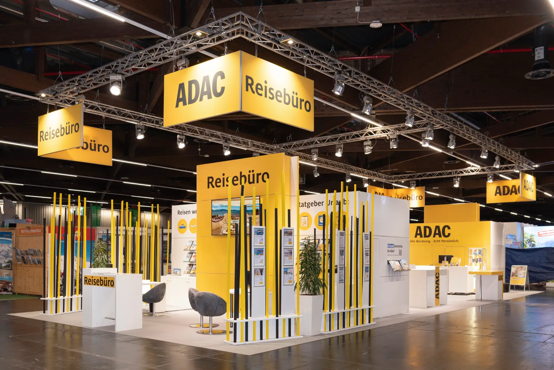 Messebau Nürnberg – ADAC Reisebüro Stand mit gelben Stehlen, zentralem „Reisebüro“-Modul, Monitor und Prospektdisplays auf der Freizeit Touristik & Garten 2025 in Nürnberg.