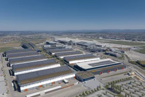 Luftaufnahme eines großen Industriekomplexes in der Nähe der Messe Stuttgart mit mehreren rechteckigen Lagerhallen, die mit Solarmodulen ausgestattet sind. Das Gelände ist von Straßen und weitläufigen Freiflächen umgeben, alles unter einem strahlend blauen Himmel.