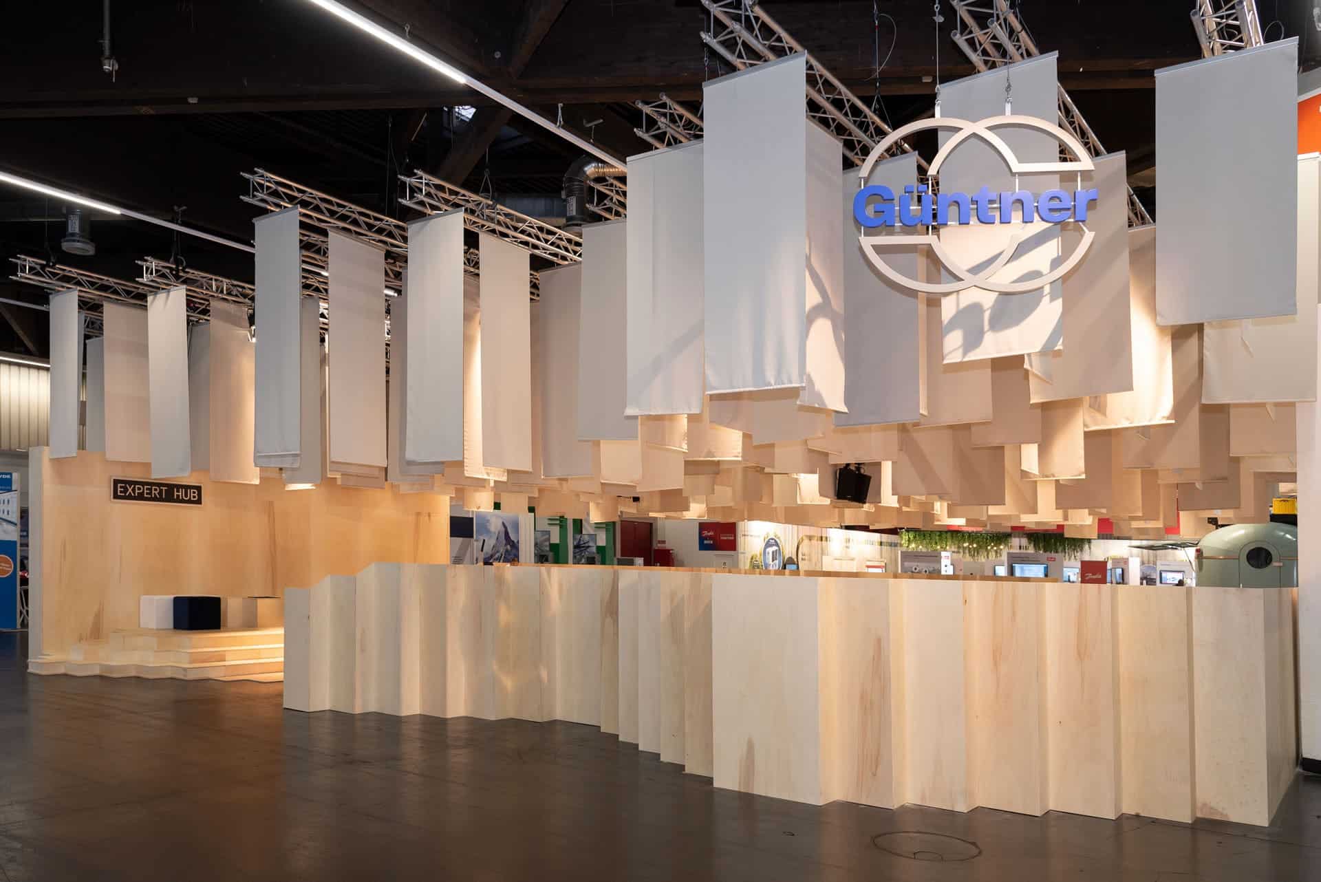 Ein innovativer Messestand mit hellen, hängenden Textilpaneelen und wellenförmigen Holzverkleidungen. Das schwebende Decken-Design, kombiniert mit einer offenen, modernen Architektur, spiegelt ein kreatives und hochwertiges Messestand Design wider. Der Expert-Hub-Bereich ist klar strukturiert und lädt zum fachlichen Austausch ein.