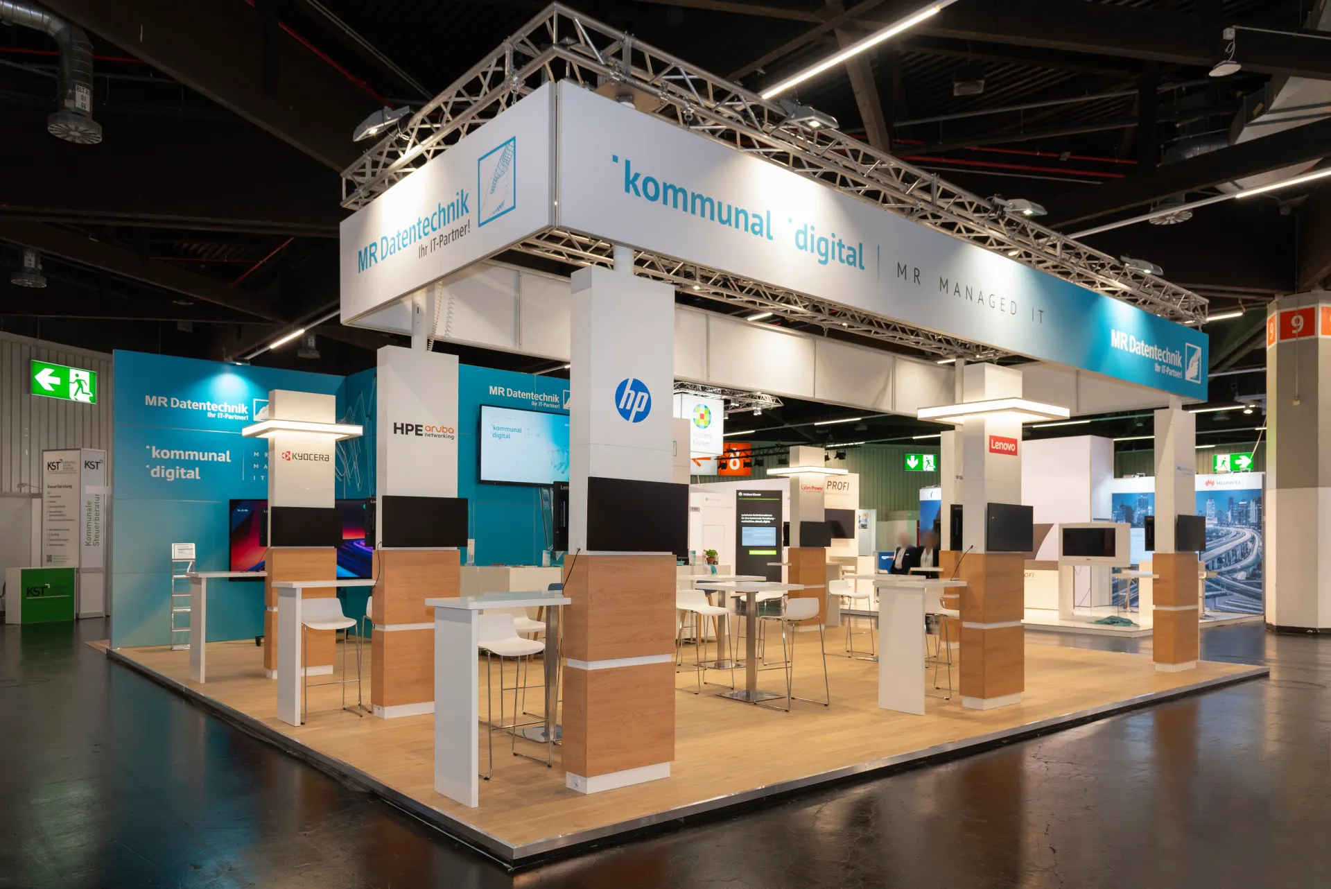 Beleuchteter Messestand von MR Datentechnik auf der Kommunale Nürnberg mit kommunal digital Banner, weißen Säulen, Holz-Stehtischen und Monitoren im Messebau.