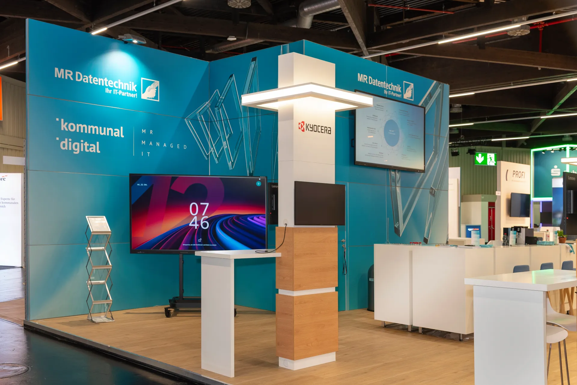 Detailansicht des MR-Datentechnik-Messestands in Nürnberg in Blau und Weiß mit Großbildschirm, Kyocera-Logo, Prospektständer und heller Holzoptik.