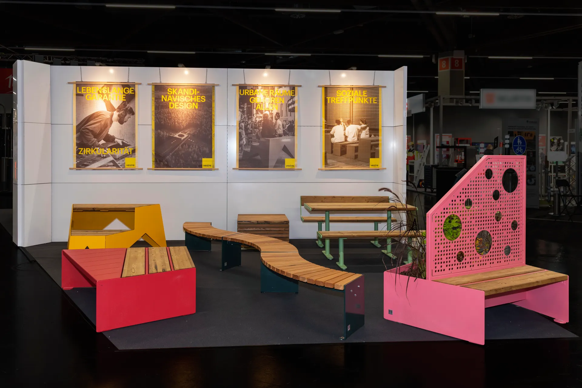 Messebau-Stand von Vestre in Nürnberg mit geschwungener Holzbank, roter und gelber Sitzmöbel, pinkfarbenem Lochblech-Element und gelben Postern an der Rückwand.
