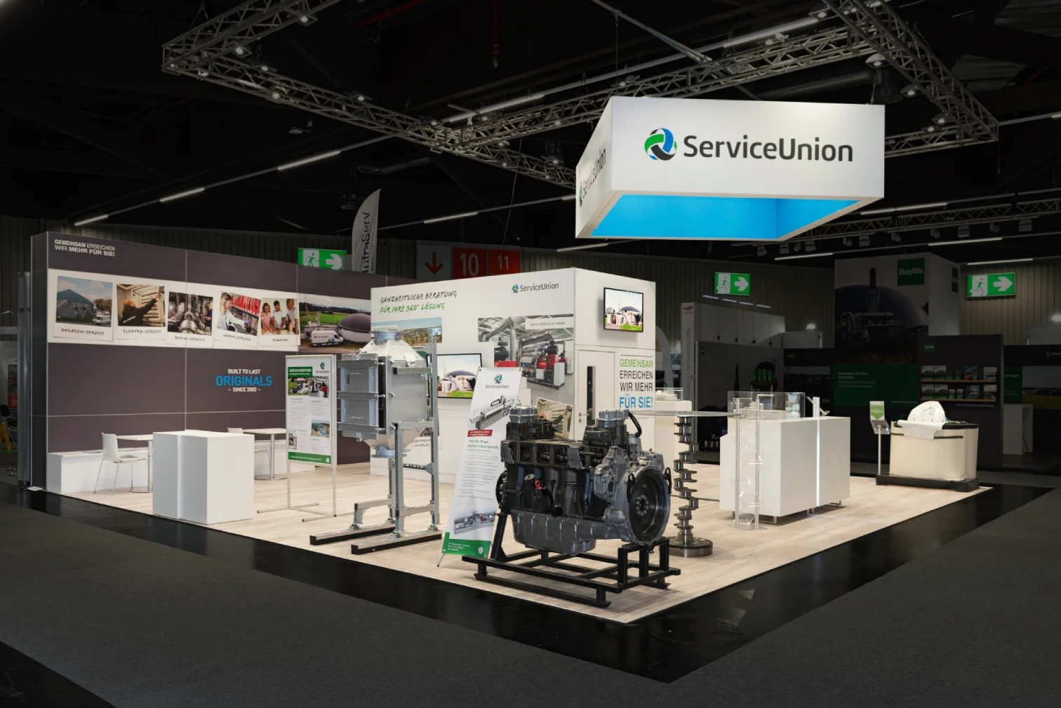 agriKomp Messestand in Nürnberg mit beleuchtetem ServiceUnion-Deckenwürfel, ausgestelltem Motorblock und Wand mit Servicefotos rund um Biogas
