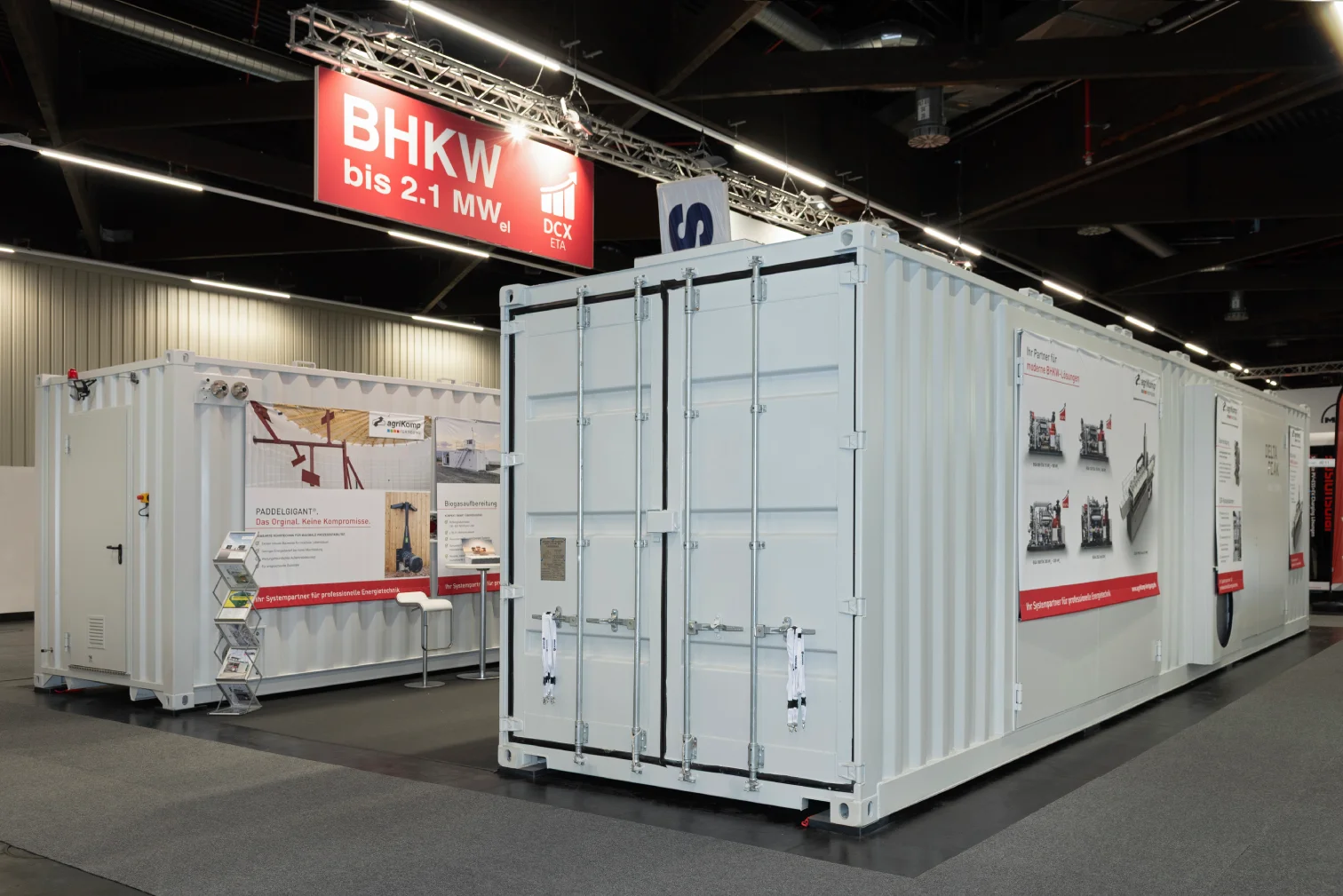 Zwei weiße Container des agriKomp-Messestands mit rotem BHKW-Banner und bedruckter Seitenwand in einer Messehalle in Nürnberg.
