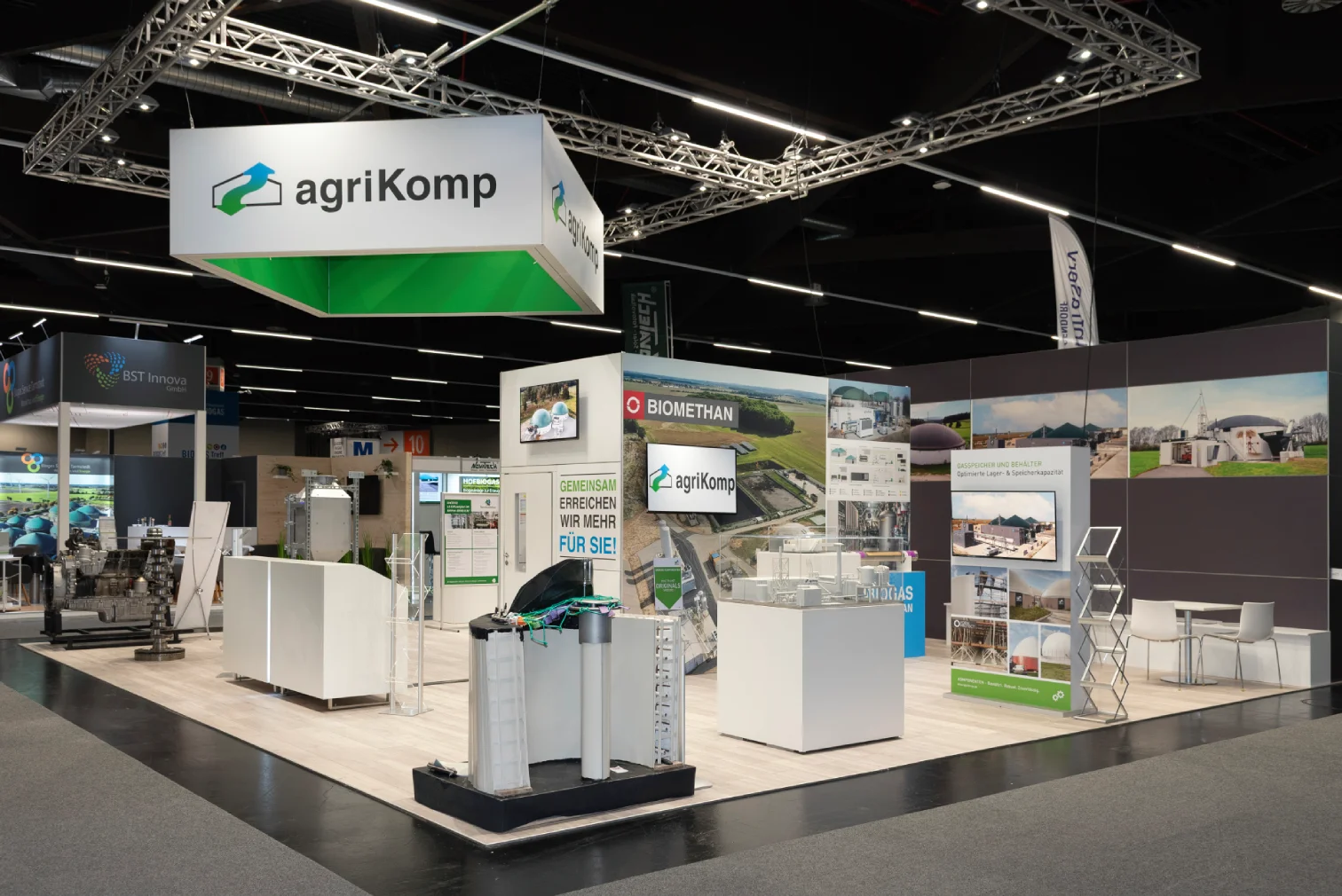 Beleuchteter agriKomp-Messestand in Nürnberg mit Logo-Banner über der Standfläche, ausgestellten Biogas-Anlagenmodellen und grüner Bildwand zum Thema Biomethan.