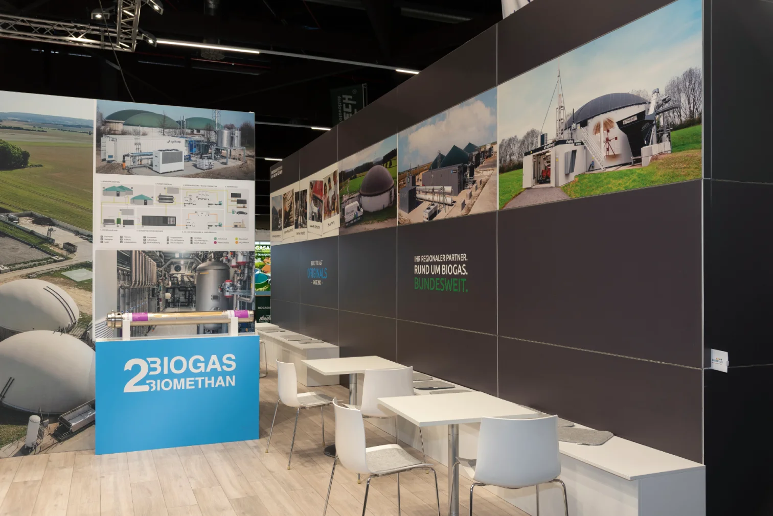 Sitzbereich am agriKomp-Messestand in Nürnberg mit weißen Tischen und Stühlen vor einer Bildwand mit Biogas- und Biomethan-Motiven und blauem 2BIOGAS BIOMETHAN-Banner.