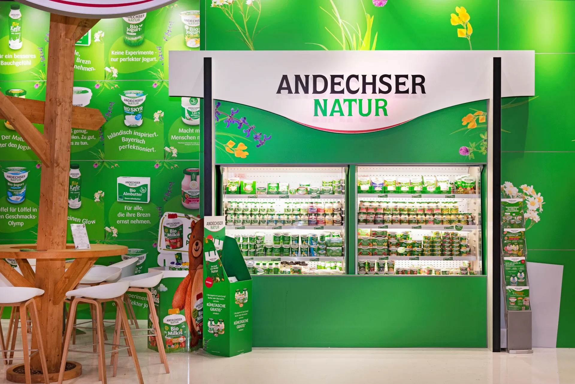 Grün gestalteter Messestand von Andechser Natur in Nürnberg mit beleuchtetem Kühlregal voller Joghurtbecher und Holz-Sitzbereich.