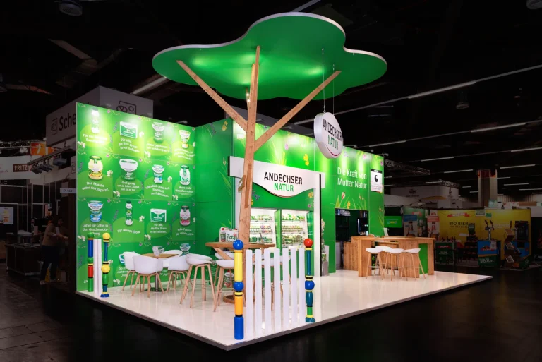 Offener Messestand von Andechser Natur in Nürnberg mit baumförmigem grünem Dachelement, grüner Markenwand und hellem Holz-Sitzbereich.