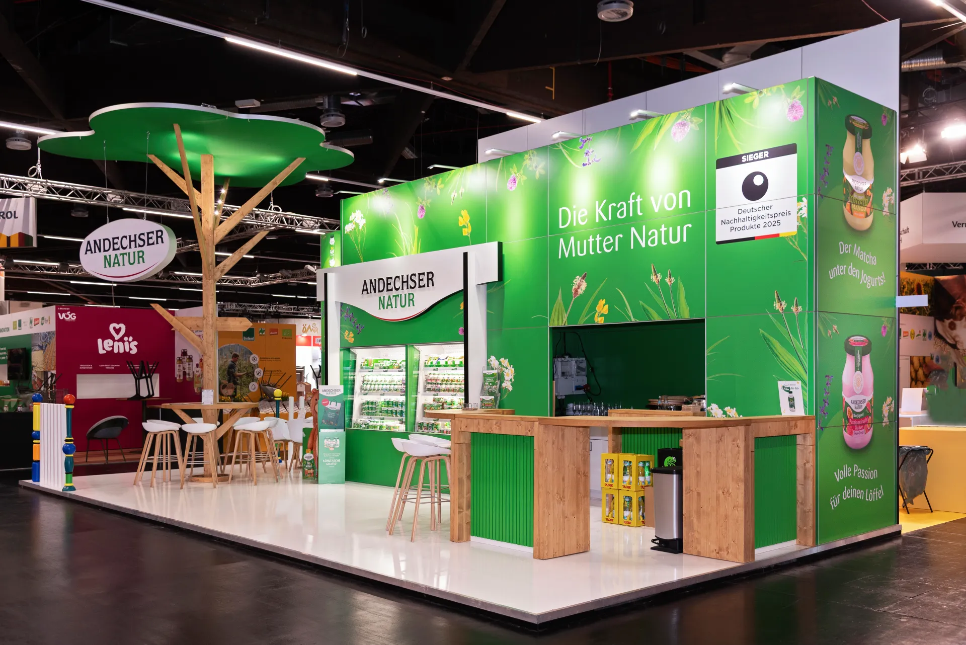 Grüner Messestand von Andechser Natur in Nürnberg mit Schriftzug Die Kraft von Mutter Natur, Theke aus Holz und baumförmigem Dachelement.
