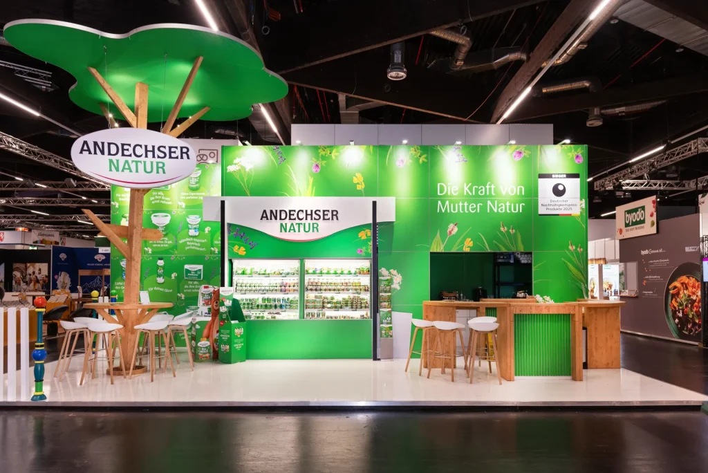 Messestand von Andechser Natur in Nürnberg mit grünem Markenbild, beleuchtetem Logo-Baum, Produktregalen und heller Holztheke mit Barhockern.