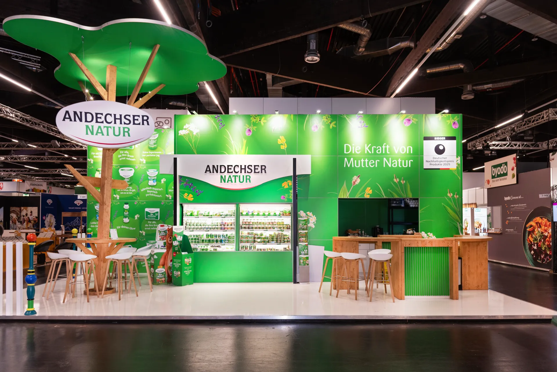 Messestand von Andechser Natur in Nürnberg mit grünem Markenbild, beleuchtetem Logo-Baum, Produktregalen und heller Holztheke mit Barhockern.