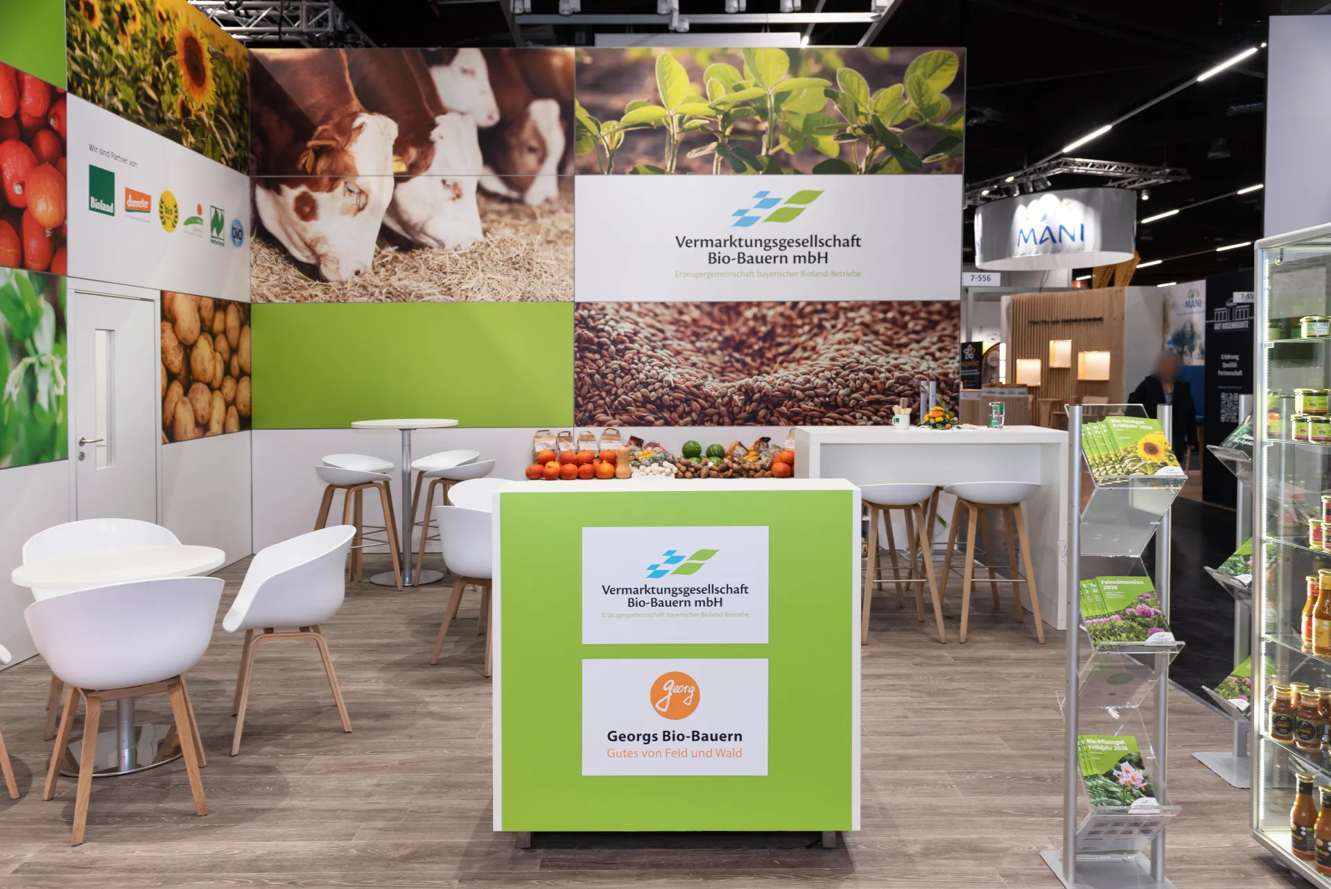 Innenraum des Bio-Bauern-Messestands in Nürnberg mit grün gebrandeter Theke „Georgs Bio-Bauern“, weißen Sitzgruppen, Holzboden und Foto-Bildwand mit Lebensmitteln.