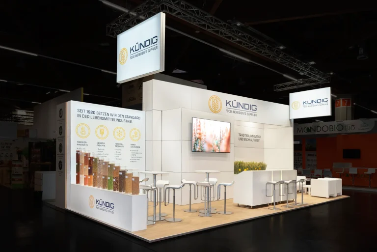 Weißer Messestand von Kündig Food Ingredients Supplier in Nürnberg mit beleuchtetem Logo-Banner, Bildschirm, Theke mit Barhockern und Rohstoffmustern.