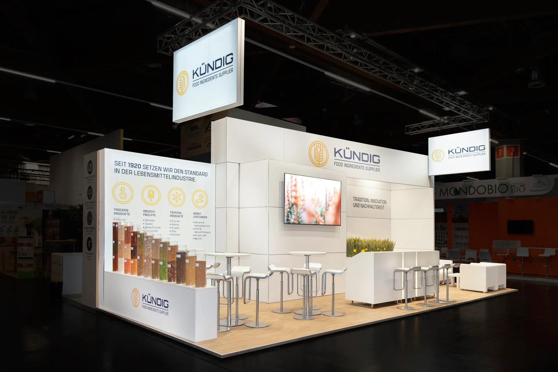 Weißer Messestand von Kündig Food Ingredients Supplier in Nürnberg mit beleuchtetem Logo-Banner, Bildschirm, Theke mit Barhockern und Rohstoffmustern.