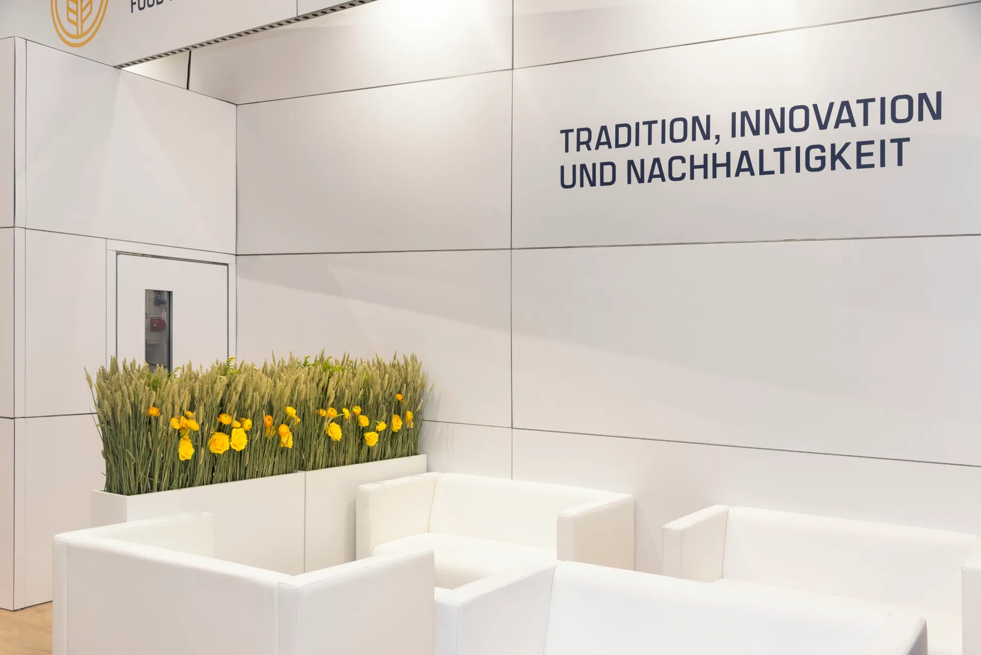 Wandausschnitt des Kündig-Messestands mit Schriftzug Tradition, Innovation und Nachhaltigkeit, Getreide-Blumen-Pflanzkasten und weißen Ledersesseln.
