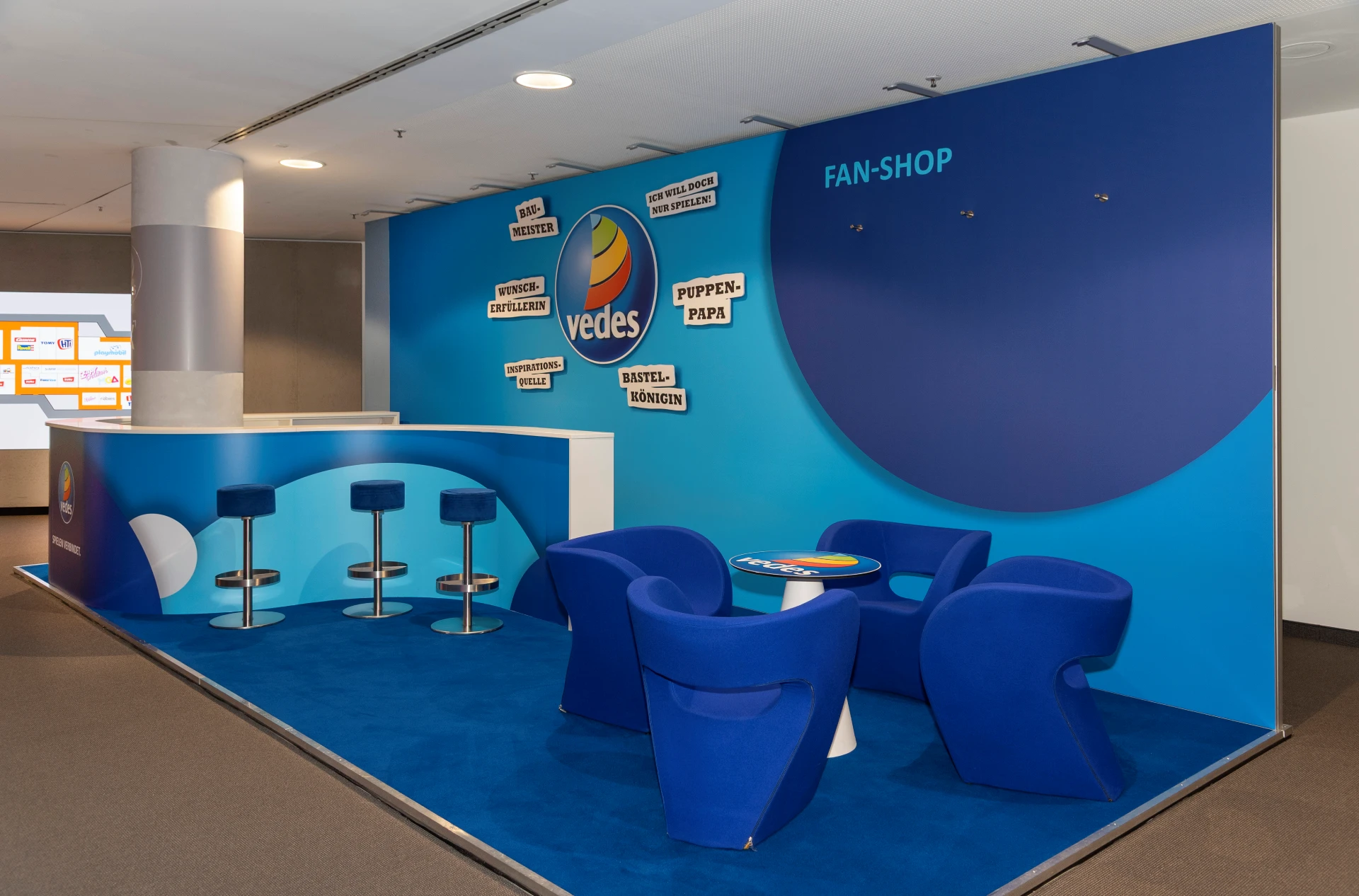 Blauer Vedes Messestand auf der Spielwarenmesse mit Logo, Bereich Fan-Shop, Theke mit Barhockern und blauen Sesseln