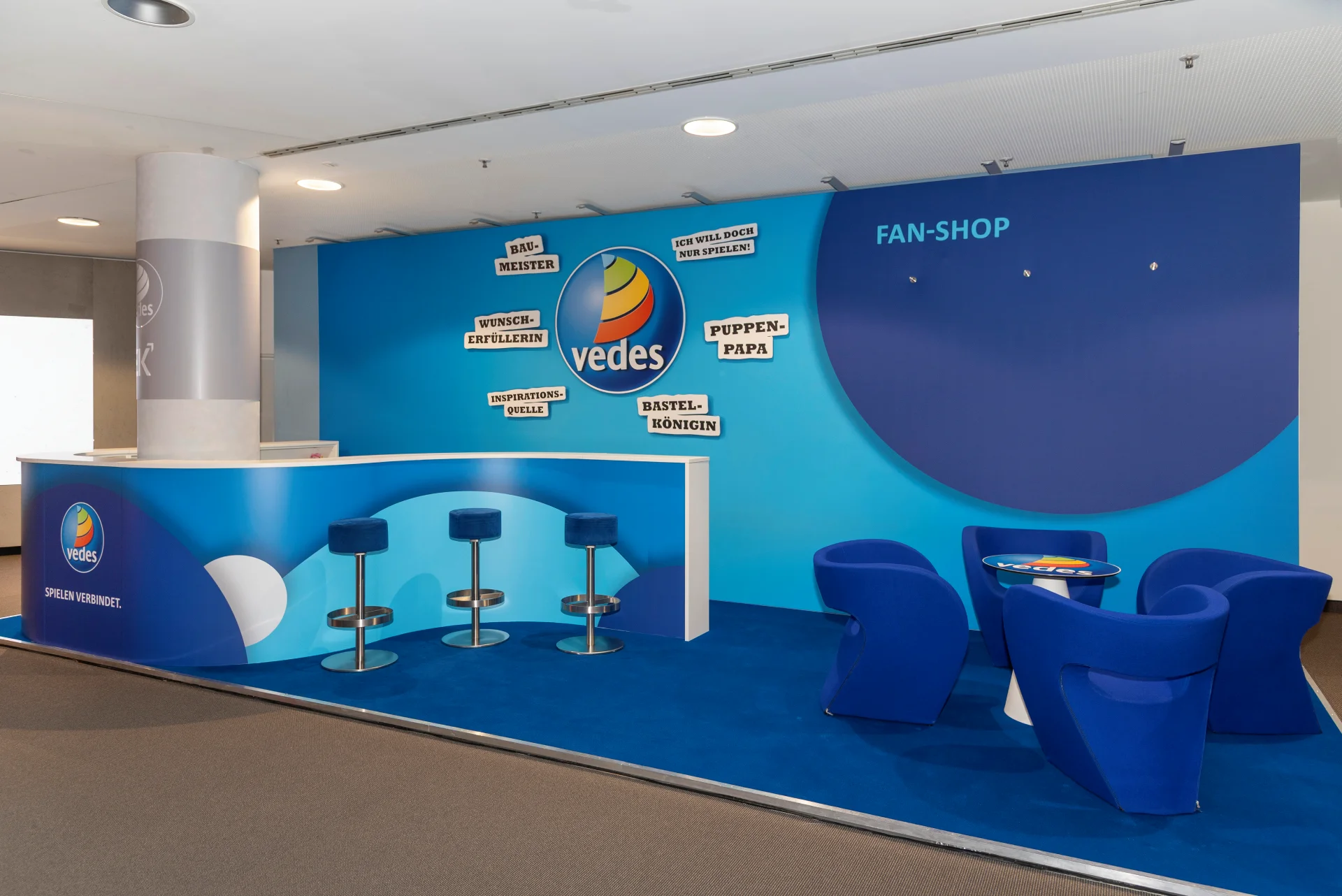 Vedes Messestand auf der Spielwarenmesse mit Logo und Slogan Spielen verbindet, Bartheke, Barhockern und blauen Loungesesseln