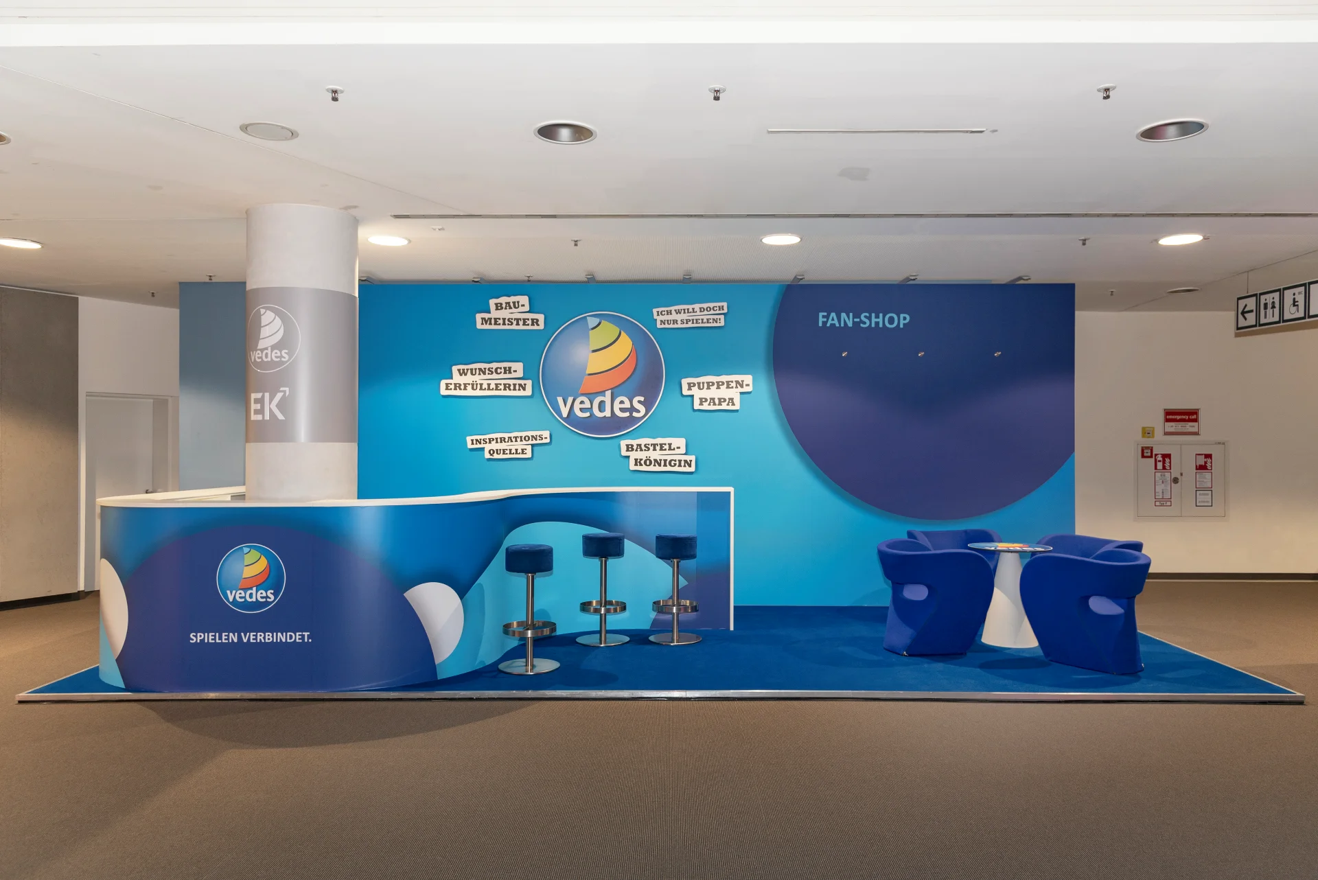 Vedes Messestand in Blau mit Logo, Slogan Spielen verbindet auf der Theke, Fan-Shop-Wand und blauer Sitzgruppe auf der Spielwarenmesse.