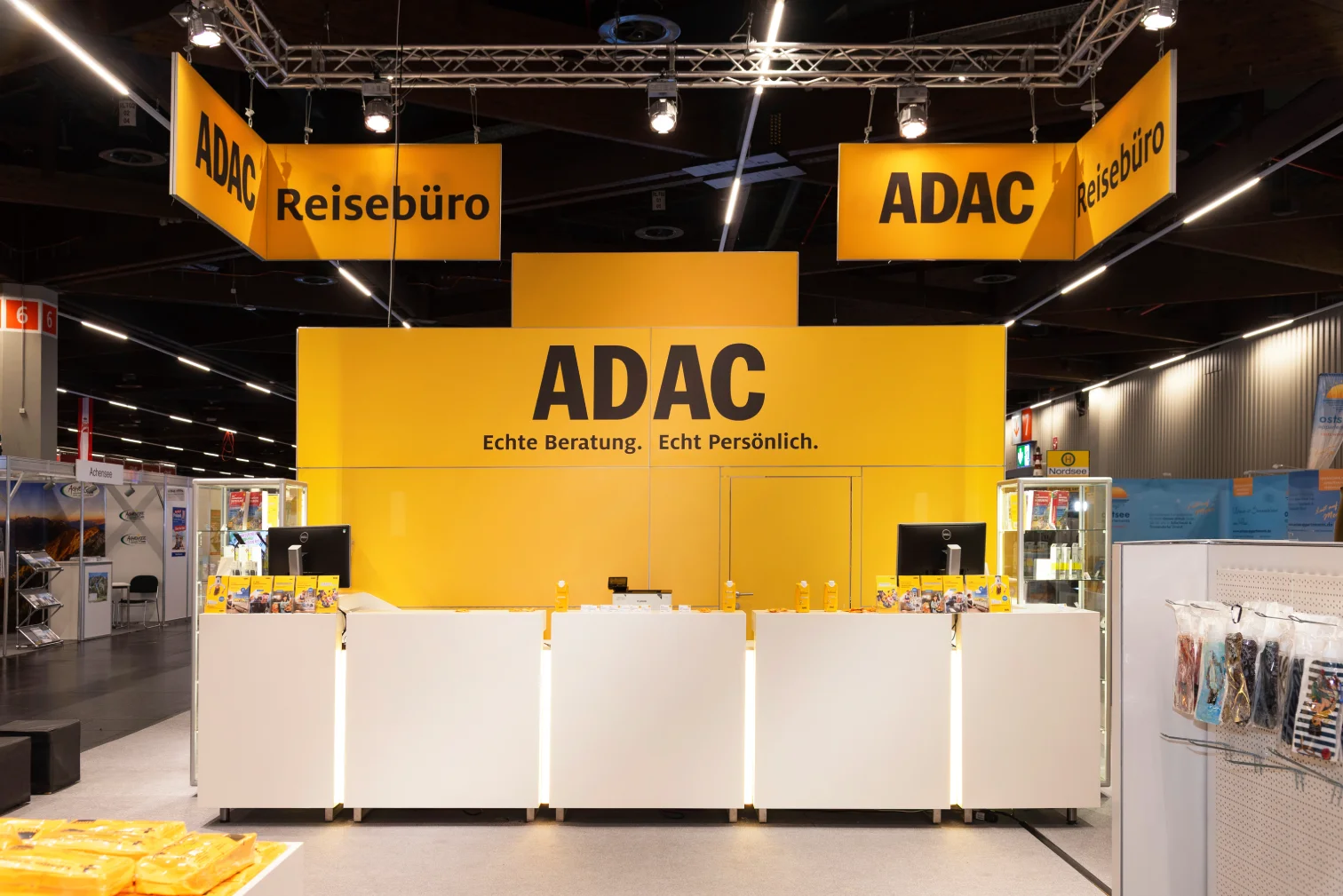 Großer ADAC-Reisebüro-Messestand auf der Freizeitmesse Nürnberg mit langer weißer Beratungstheke, gelber Rückwand und hängenden ADAC-Reisebüro-Schildern.