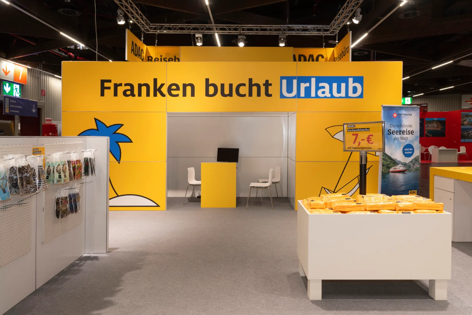 Gelber ADAC-Messestand in Nürnberg mit Durchgang, Schriftzug Franken bucht Urlaub, Sitzbereich, Prospektständern und Reise-Angebotsschild.