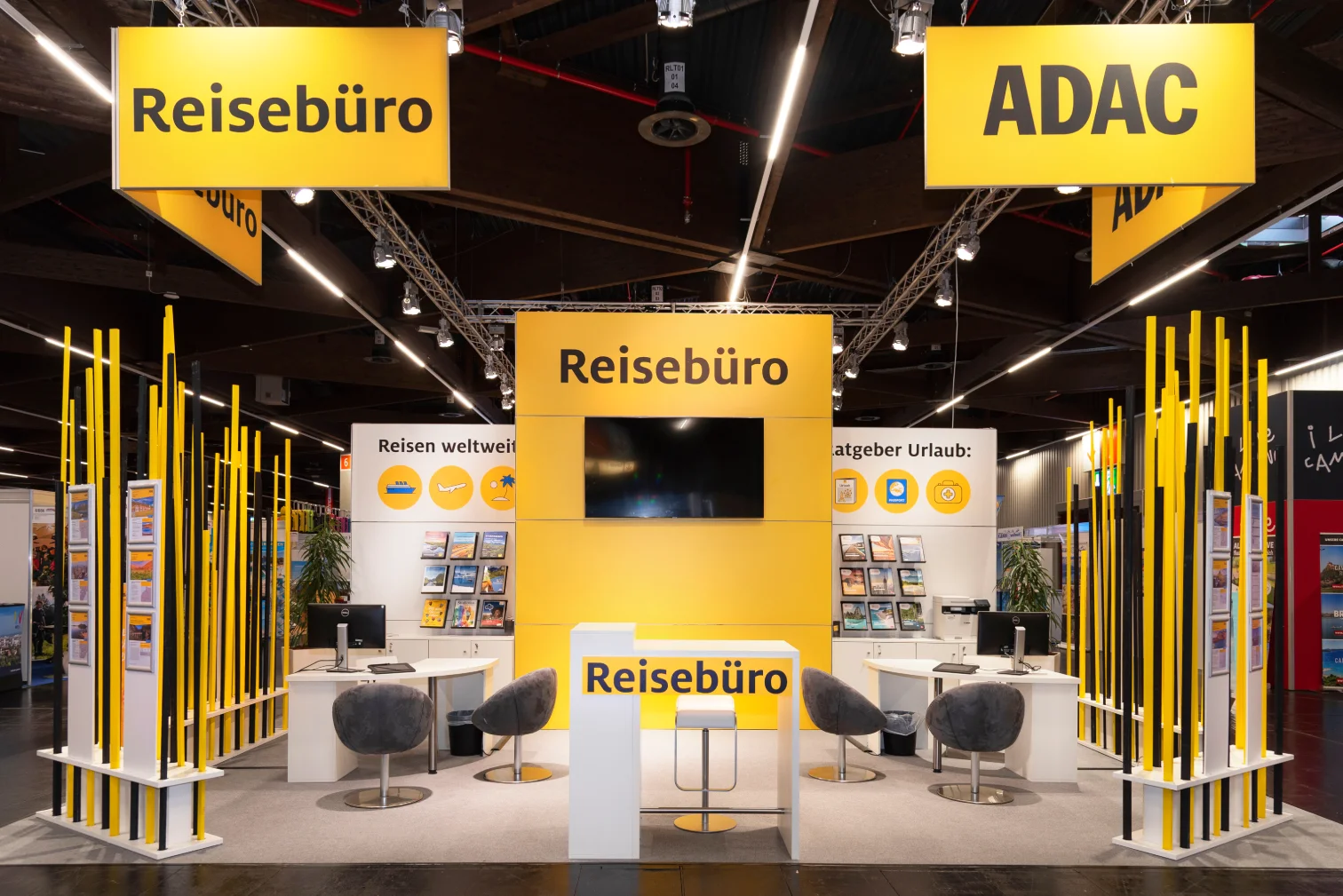 ADAC-Reisebüro-Messestand auf der Freizeitmesse Nürnberg mit gelben Deckenbannern, Bildschirmwand, Prospektregalen und Sitzgruppen mit grauen Sesseln.