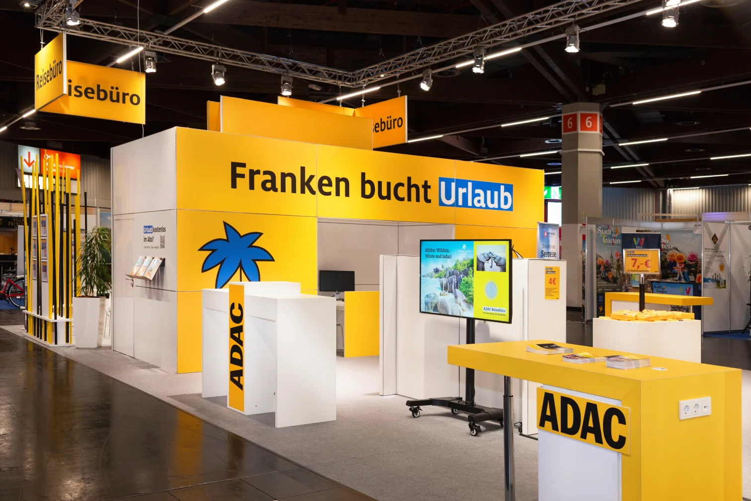 ADAC-Messestand auf der Freizeitmesse Nürnberg mit Schriftzug Franken bucht Urlaub, blauem Palmen-Logo, Monitor und gelben Theken.