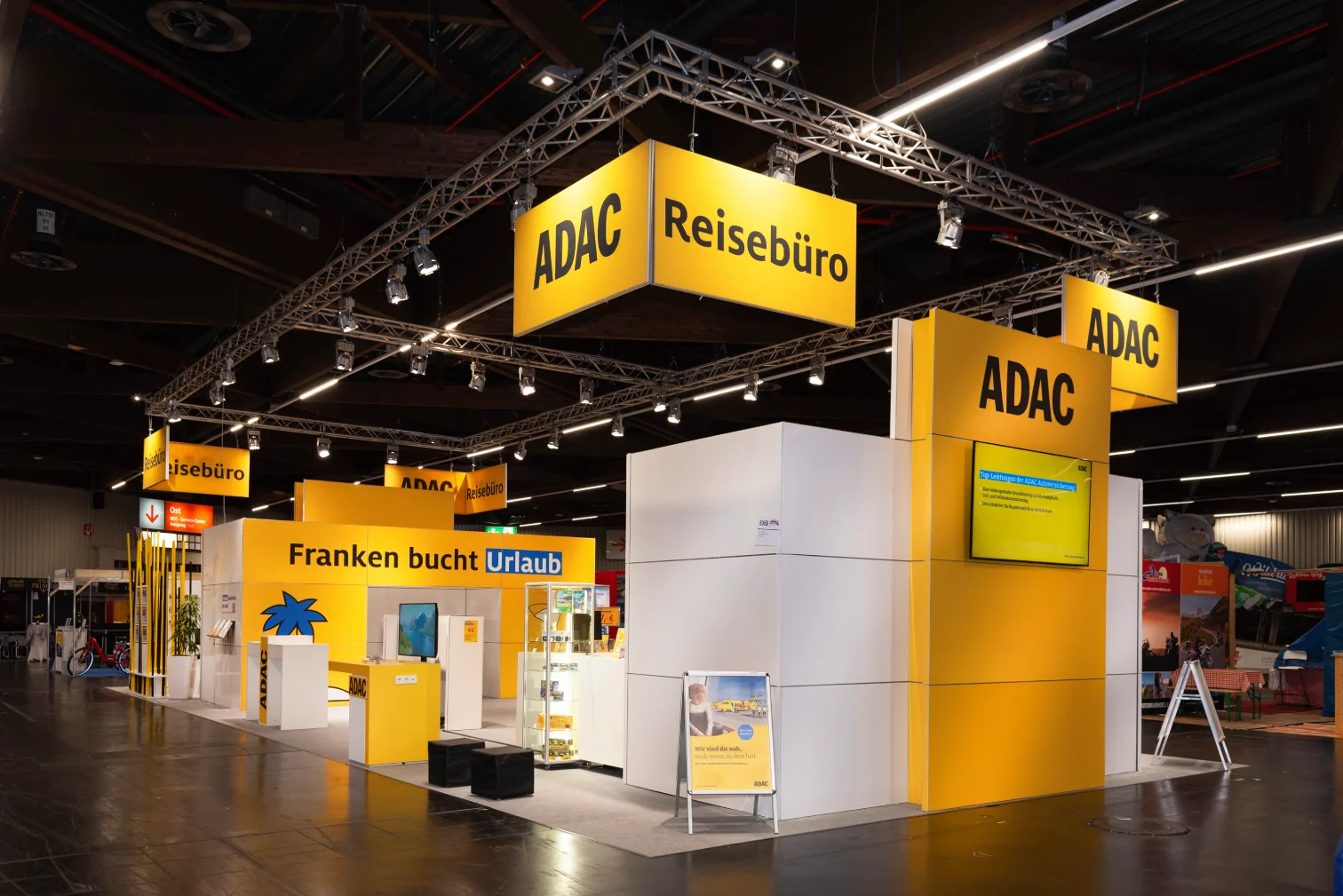 Weiß-gelber ADAC Reisebüro-Messestand auf der Freizeitmesse Nürnberg mit beleuchtetem Deckenbanner über Traversen und Tresen mit Slogan Franken bucht Urlaub.