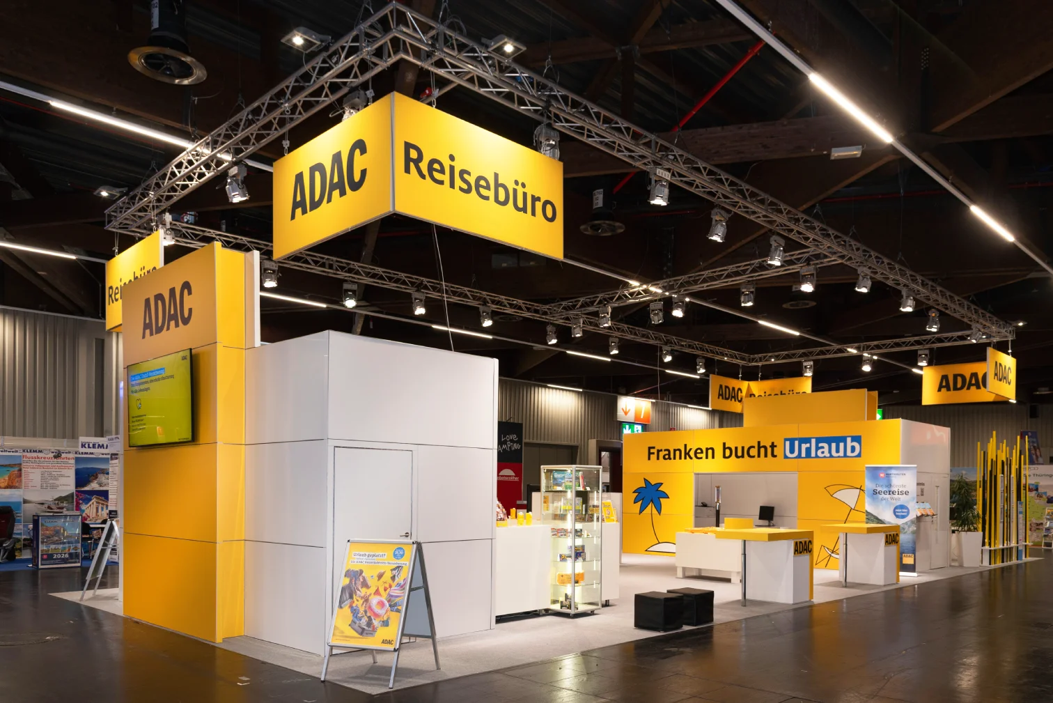 Großer ADAC Reisebüro-Messestand auf der Freizeitmesse Nürnberg mit gelben Leuchtbannern an Deckentraversen, weißer Kabine und Theken mit Franken bucht Urlaub.
