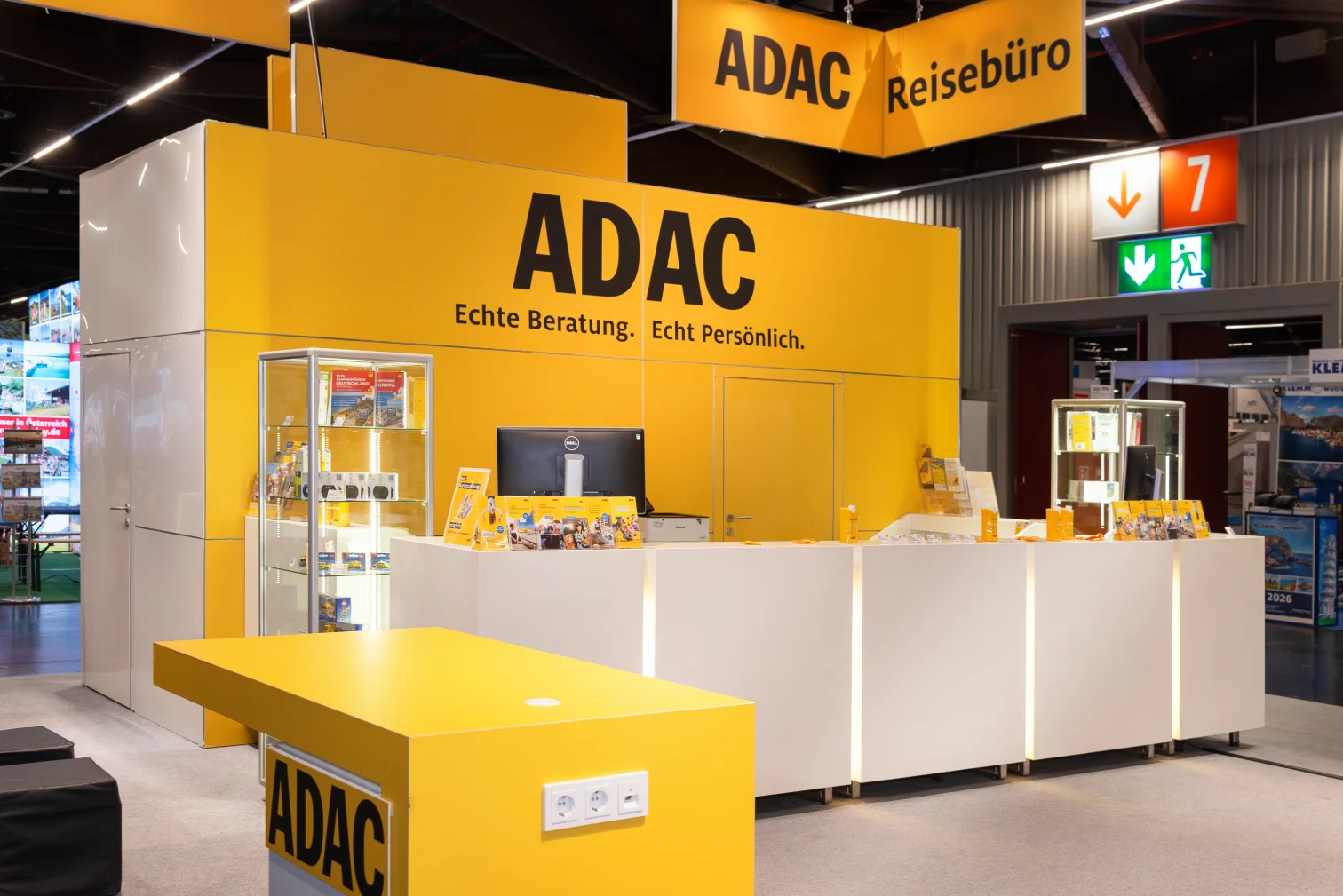 ADAC-Reisebüro-Messestand in Nürnberg mit gelber Theke, Schriftzug ADAC Echte Beratung. Echt Persönlich. und beleuchteter Glasvitrine.