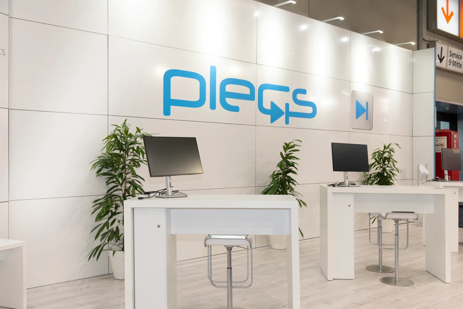 Weißer Messestand mit blauem Plecs-Logo an der Rückwand, zwei Stehtischen mit Monitoren und Grünpflanzen, Messebau für Plexim auf der PCIM Nürnberg.