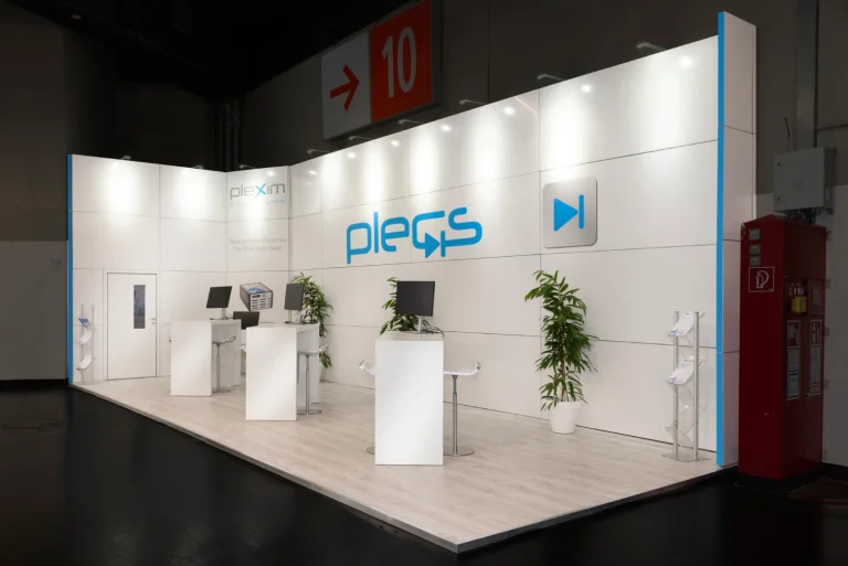 Weißer Plexim-Messestand mit Plecs-Logo, Beratungstheken und Grünpflanzen im Gang neben Hallenschild 10 auf der PCIM Nürnberg, Messebau.
