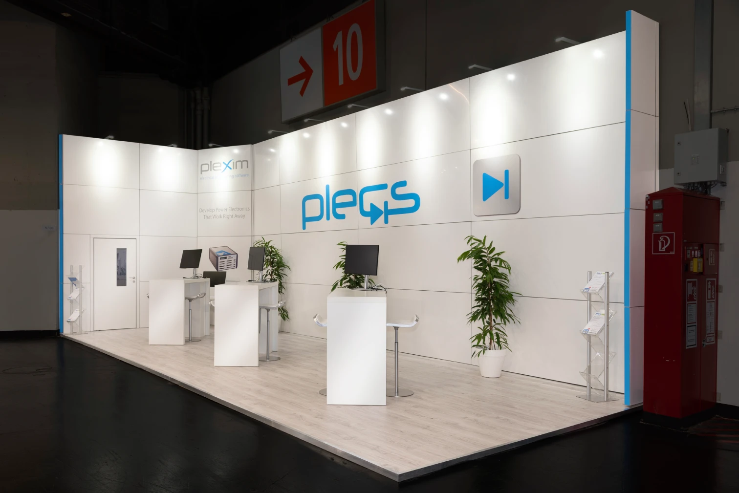 Weißer Plexim-Messestand mit Plecs-Logo, Beratungstheken und Grünpflanzen im Gang neben Hallenschild 10 auf der PCIM Nürnberg, Messebau.