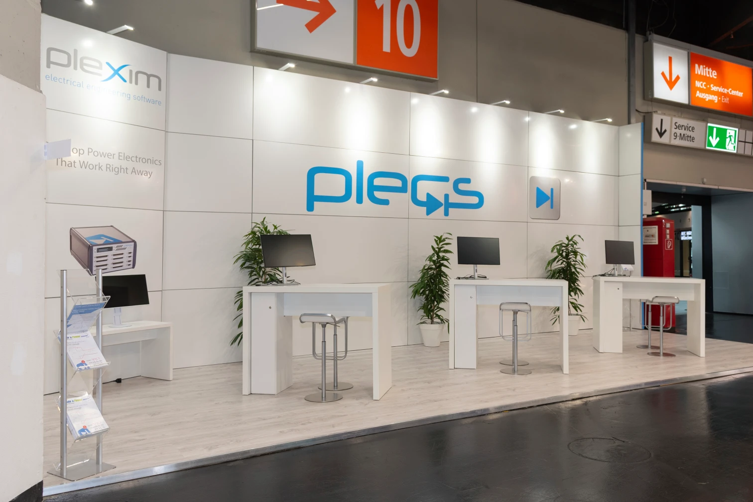 Messebau-Stand von Plexim auf der PCIM Nuernberg mit drei weissen Beratungstischen, Monitoren und Pflanzen vor der blauen PLECS-Wand.